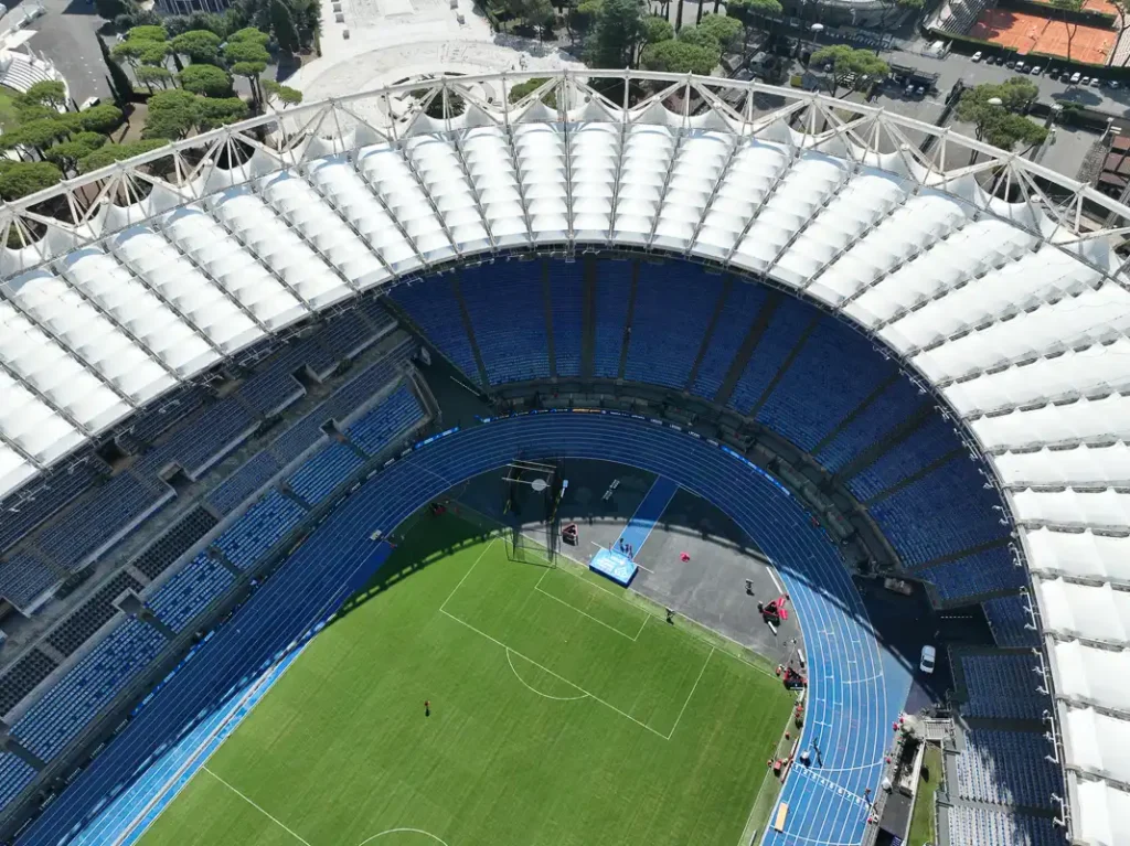 Sfotografowany z drona w samo południe Stadio Olimpico, z silnym kontrastem między białym zadaszeniem a głębokim cieniem rzucanym na trybuny