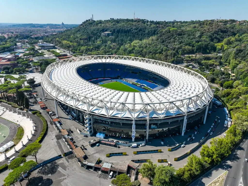 Panoramiczne ujęcie z drona przedstawiające bryłę Stadio Olimpico na tle rzymskiej zabudowy pod bezchmurnym, błękitnym niebem.