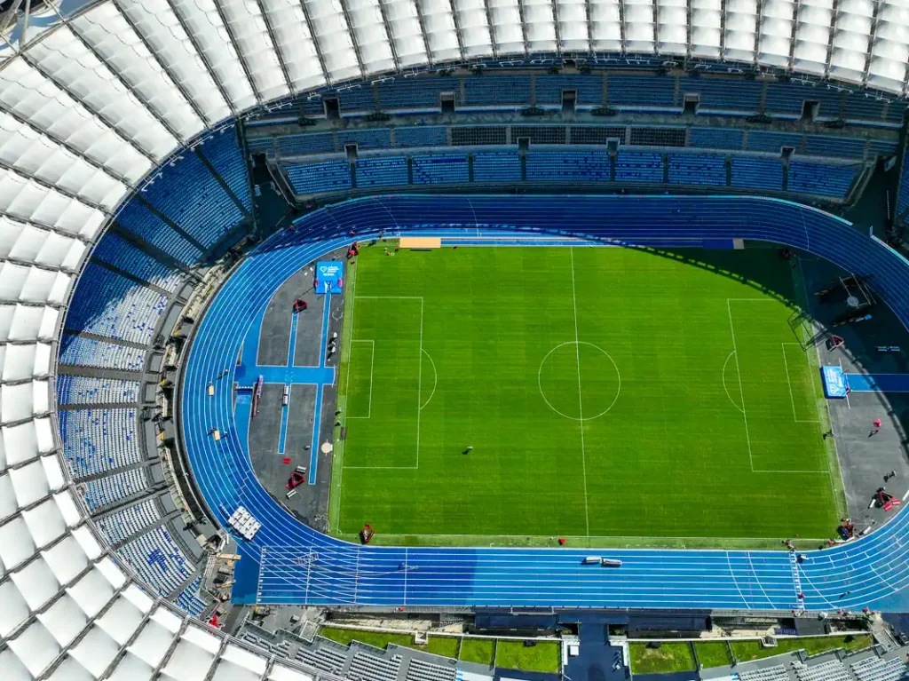 Ostre cienie rzucane przez konstrukcję dachu na błękitne sektory Stadio Olimpico podczas bezchmurnego południa.